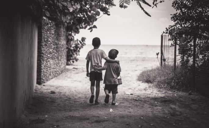 Black and white image of two boys walking down the road with fishing rods, one arm over the other.