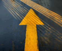 A bold yellow arrow painted on wet asphalt, pointing forward with streaks of light and tire marks crossing it, symbolizing direction, momentum, or moving ahead despite obstacles.