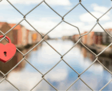 A red heart-shaped padlock attached to a chain-link fence overlooking a calm river lined with colorful buildings, symbolizing love, commitment, or emotional connection.