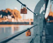 Two brass padlocks fastened to steel cables on a bridge, with a blurred cityscape and river in the background, captured in warm, golden-hour lighting.