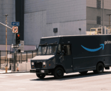 A dark Amazon Prime delivery truck drives through a city intersection under a red light.